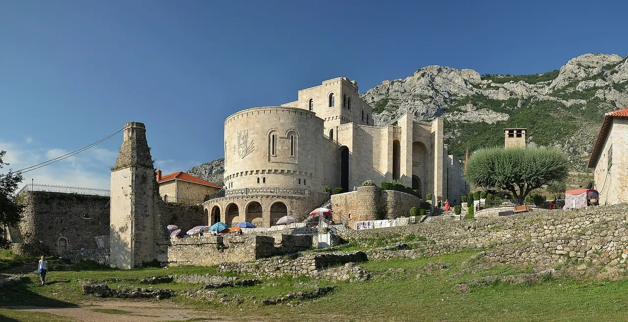 A wide view of Kruja Castle grounds: the Skanderbeg Museum tower, ruined stone walls and archaeological foundations, with the grey wall of Mount Krujë behind