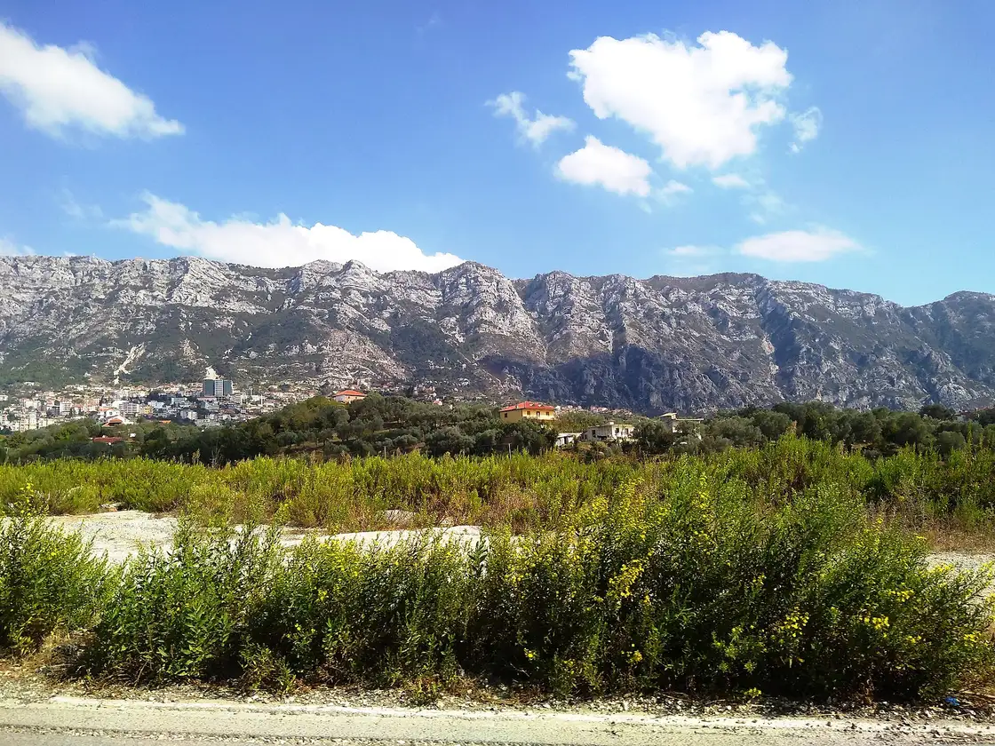 The bare grey wall of Mount Krujë rising behind the town, seen across scrubland from the approach road