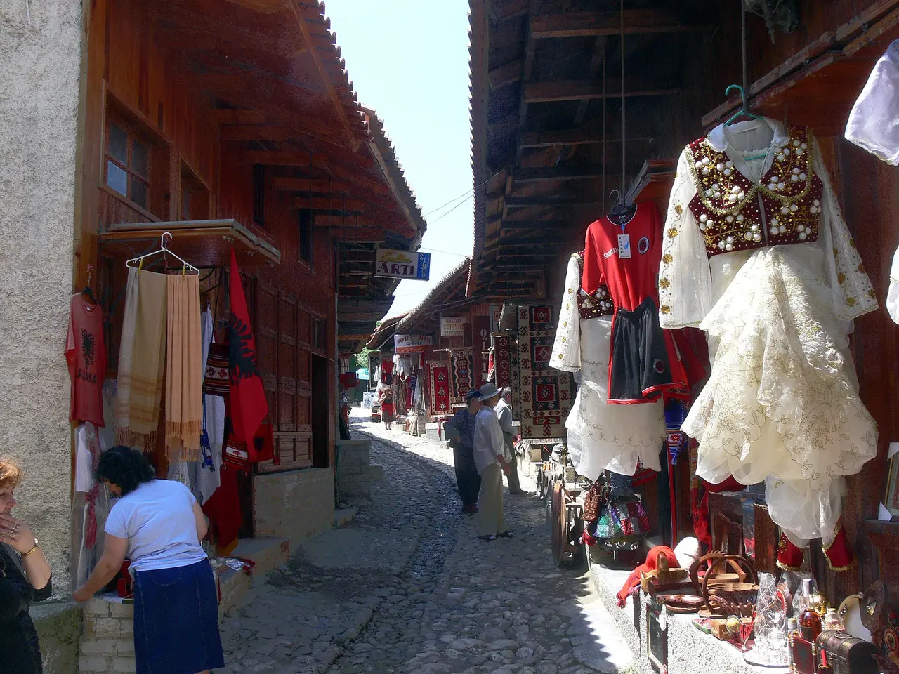 The cobbled Old Bazaar street in Kruja lined with wooden shops, hung with Albanian flags, red garments, traditional embroidered costumes and carpets, with a few shoppers walking through