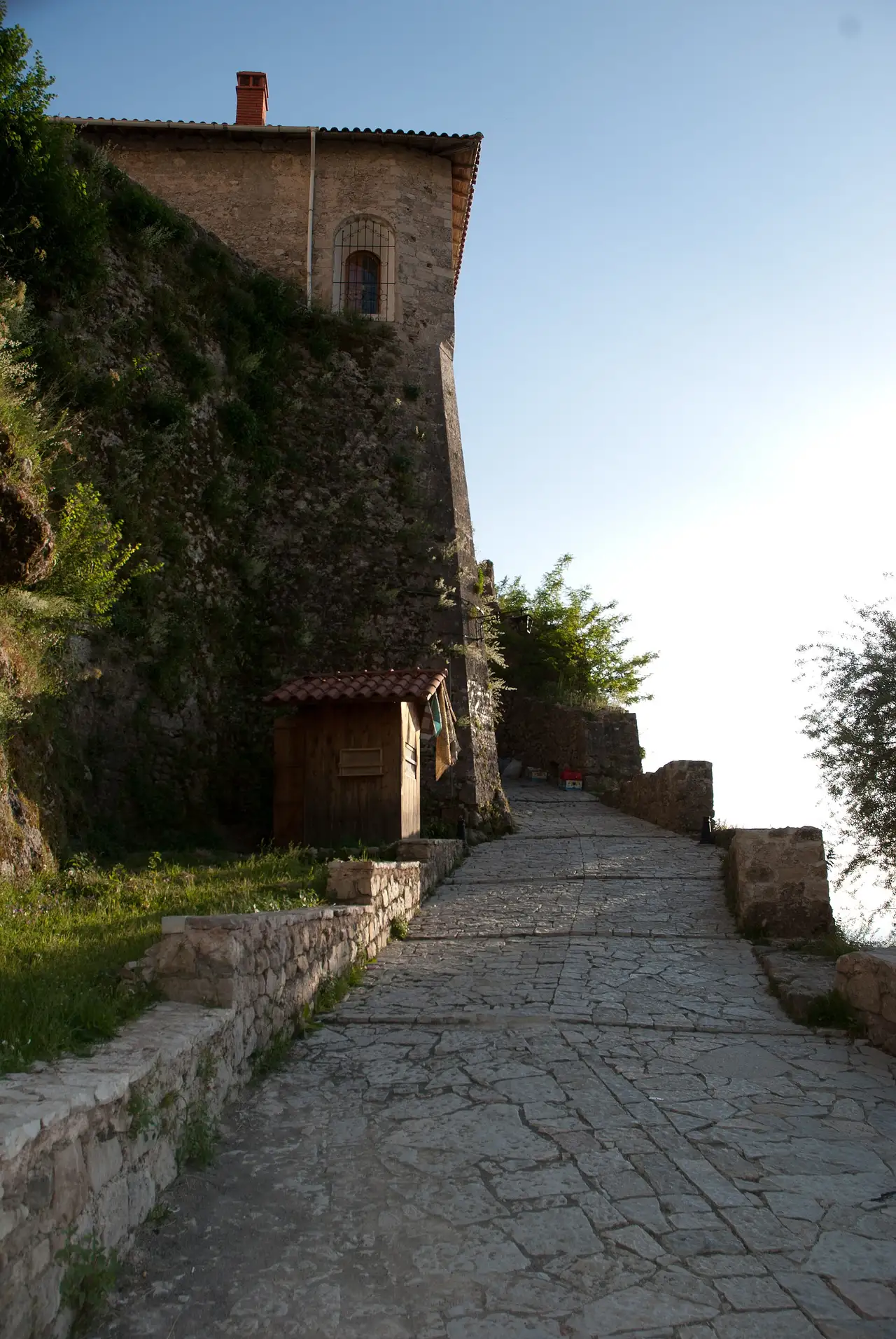 A steep flagstone lane climbing to the castle wall in Kruja, past a stone building and a small wooden stall