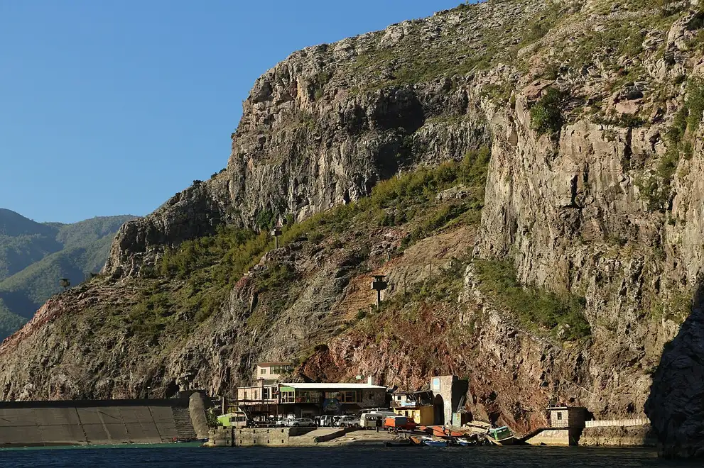 The ferry landing at Koman with vehicles and small boats gathered on a quay beneath a huge rock face