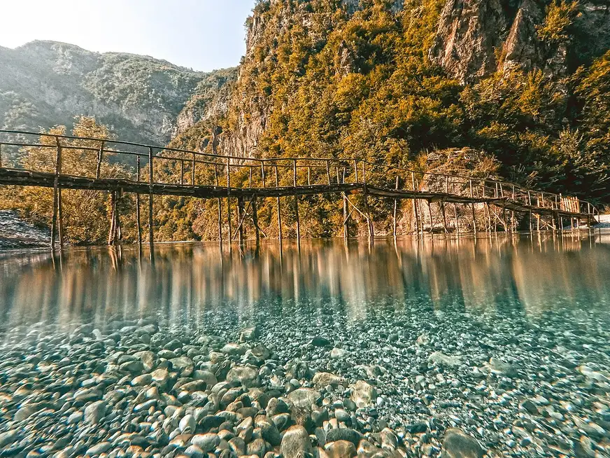 A rustic wooden footbridge over the crystal-clear turquoise water of the Shala river above Lake Koman in the Albanian Alps