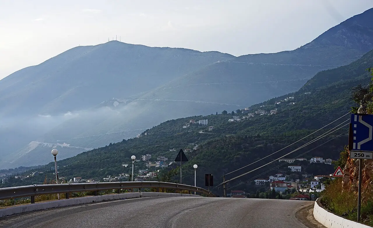 The SH8 coast road curving along a green mountainside above a hazy Riviera town on the Vlora side of the Llogara Pass
