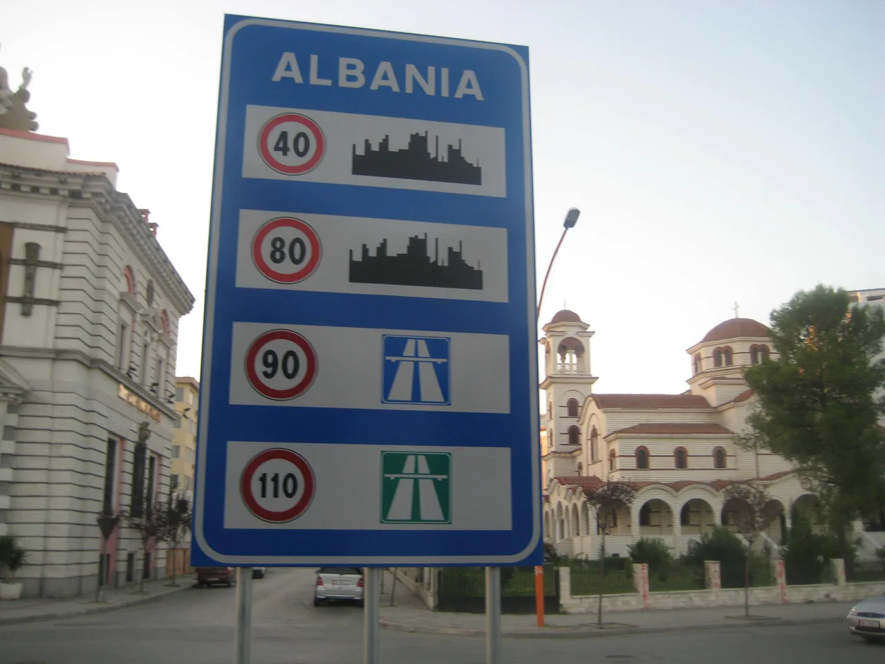 A blue road sign headed ALBANIA listing speed limits of 40, 80, 90 and 110 km/h, with an Orthodox church and a cream-coloured building behind it