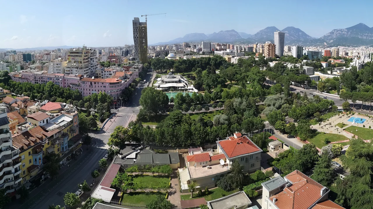 Tirana from a high-rise on a sunny day: apartment blocks and red-roofed villas around a leafy park with a pool, a tree-lined boulevard, towers and a crane, and mountains along the skyline