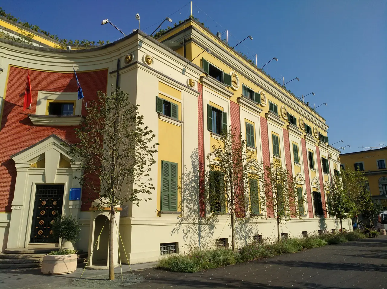 The red, yellow and cream front of the Tirana Municipality building, with green shutters and young trees along the pavement