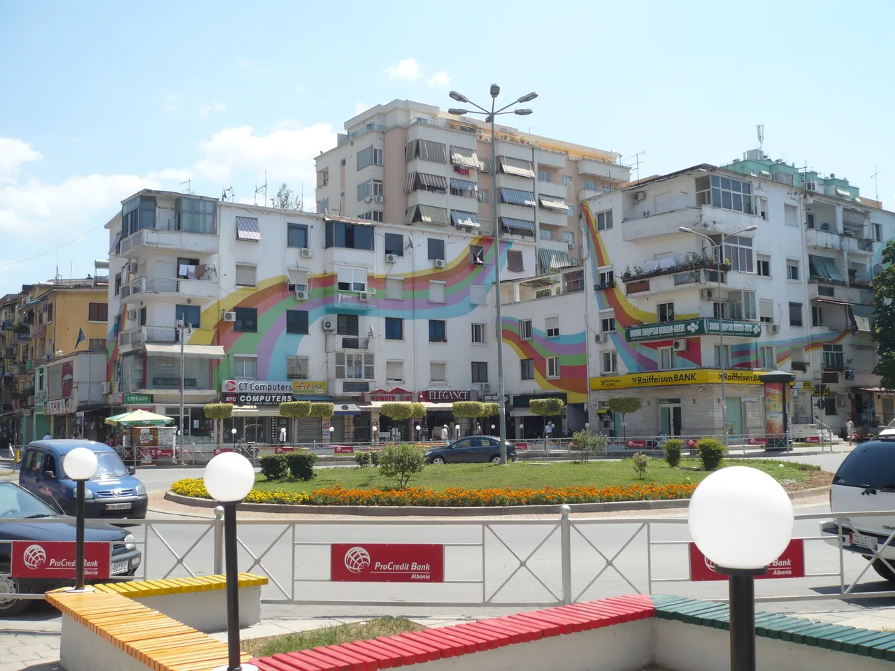 Apartment blocks painted with rainbow stripes above shops and a Raiffeisen Bank branch on a Tirana roundabout, with ProCredit Bank banners on the railing in front