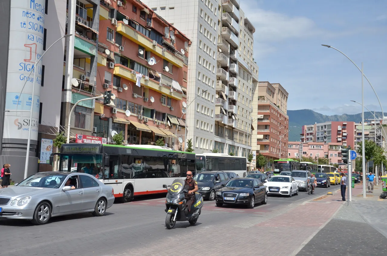City buses, cars and a scooter on a Tirana boulevard lined with pink, yellow and white apartment blocks, with mountains in the distance