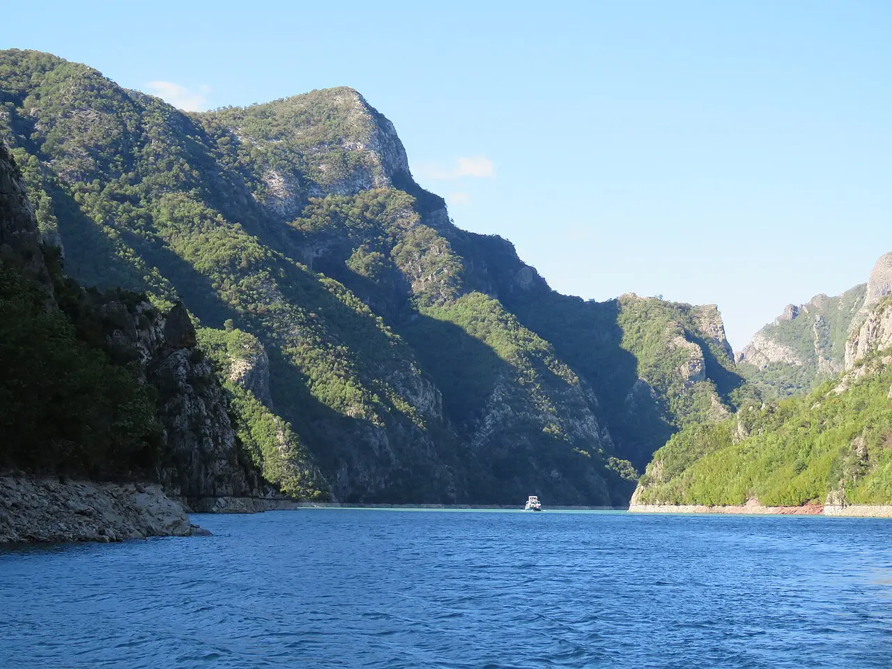 The steep forested cliffs of the Koman gorge dropping straight into blue water with a small boat