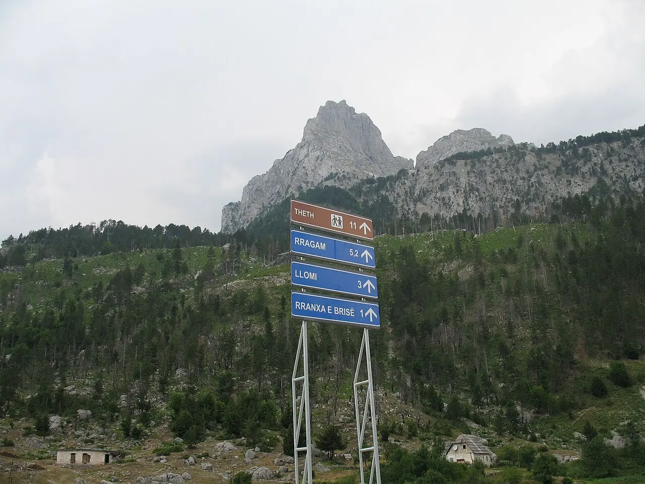 A trail signpost pointing to Theth 11 km under the jagged grey peaks of the Accursed Mountains