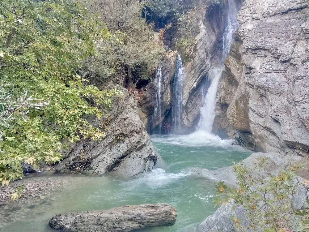 Bogovë waterfall dropping in two strands over grey rock into a green pool surrounded by trees