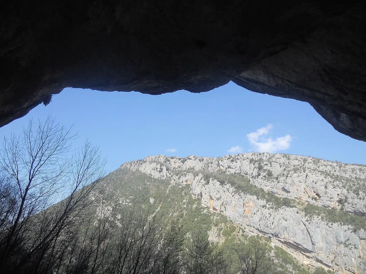 Looking out from inside the cave mouth: a dark rock arch frames blue sky and the pale limestone wall of the gorge, with bare trees in front