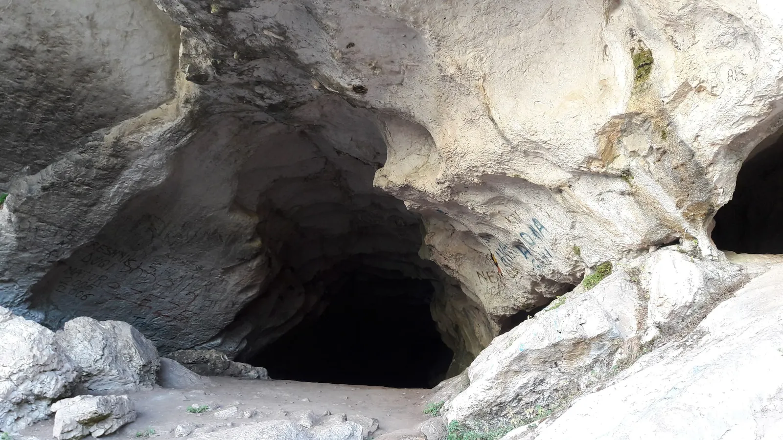 The wide, dark mouth of the Pëllumbas Black Cave in pale limestone, with a second smaller opening on the right