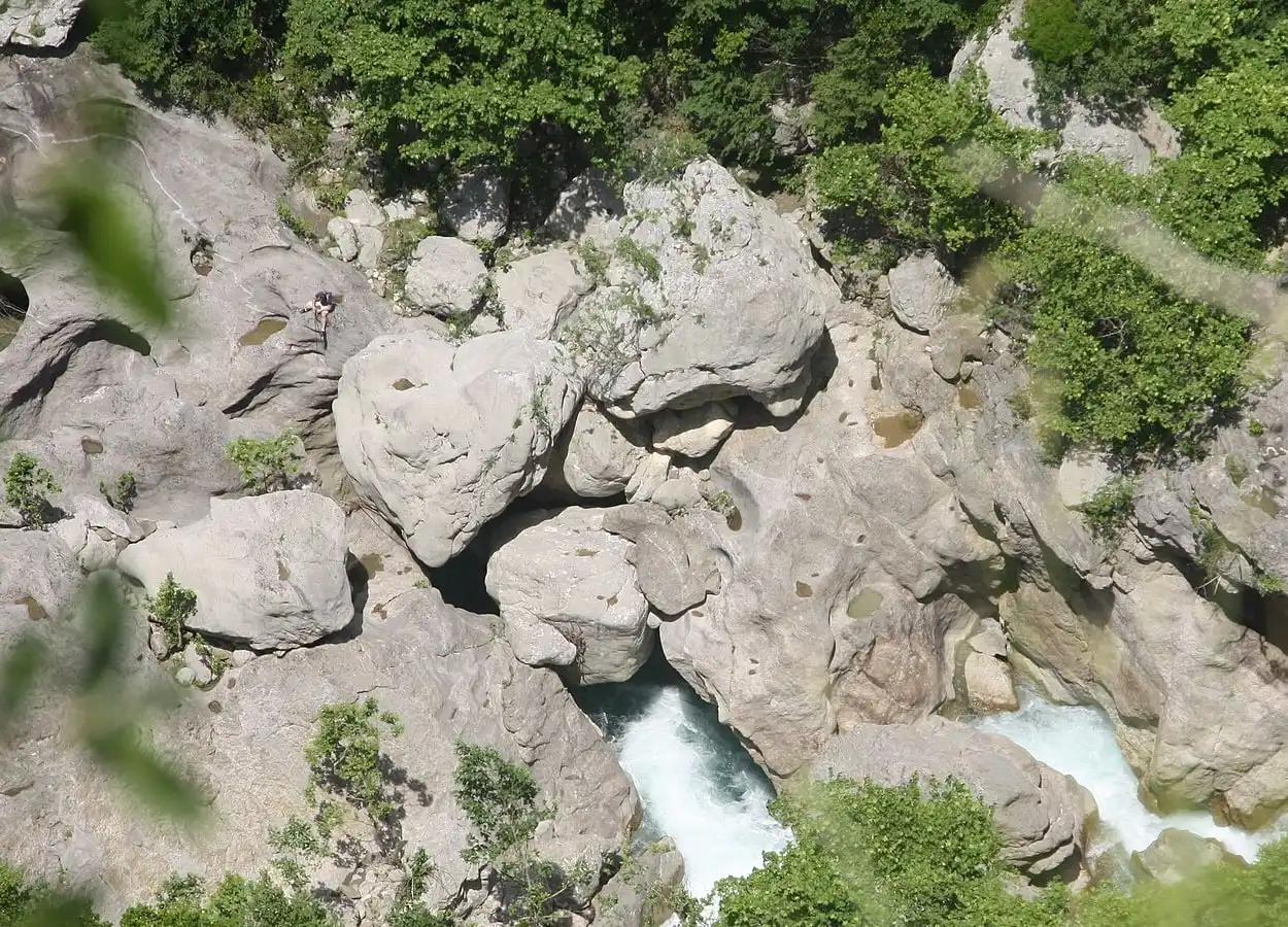 Looking down into a rocky gorge where white water runs between pale boulders, with a small figure on the rock at left