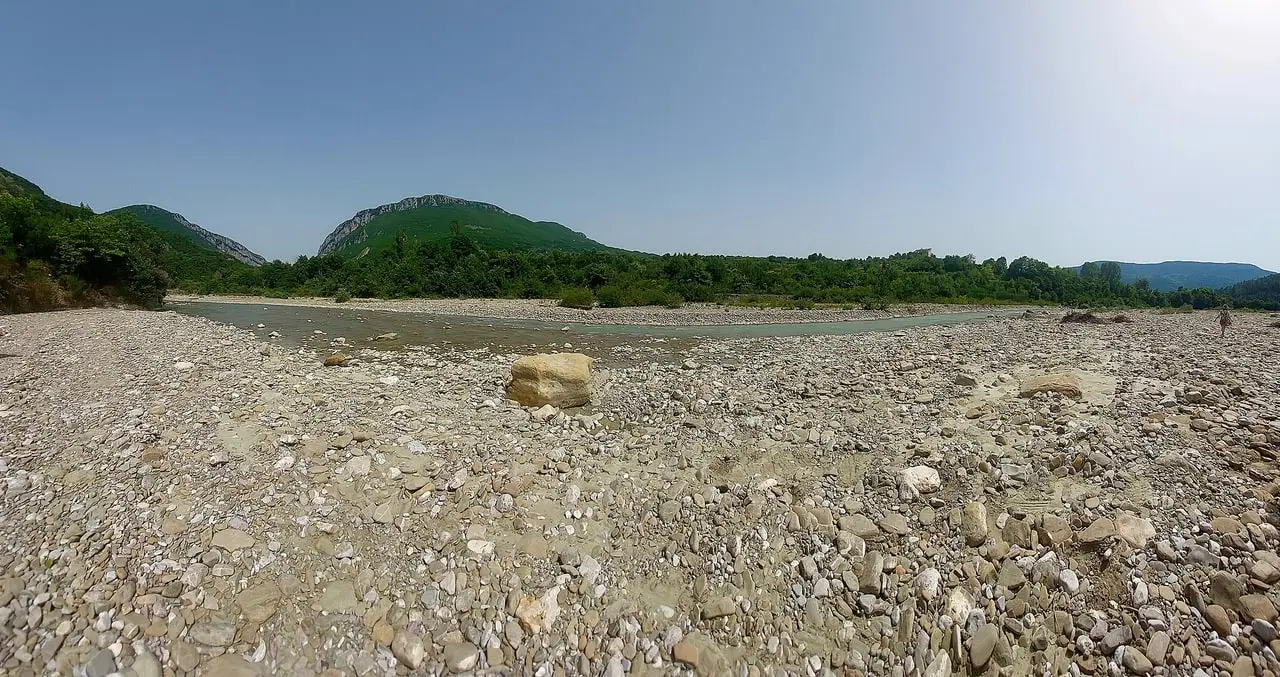 A wide gravel river bed with one shallow channel of water, a pale boulder and green wooded hills under a hazy sky