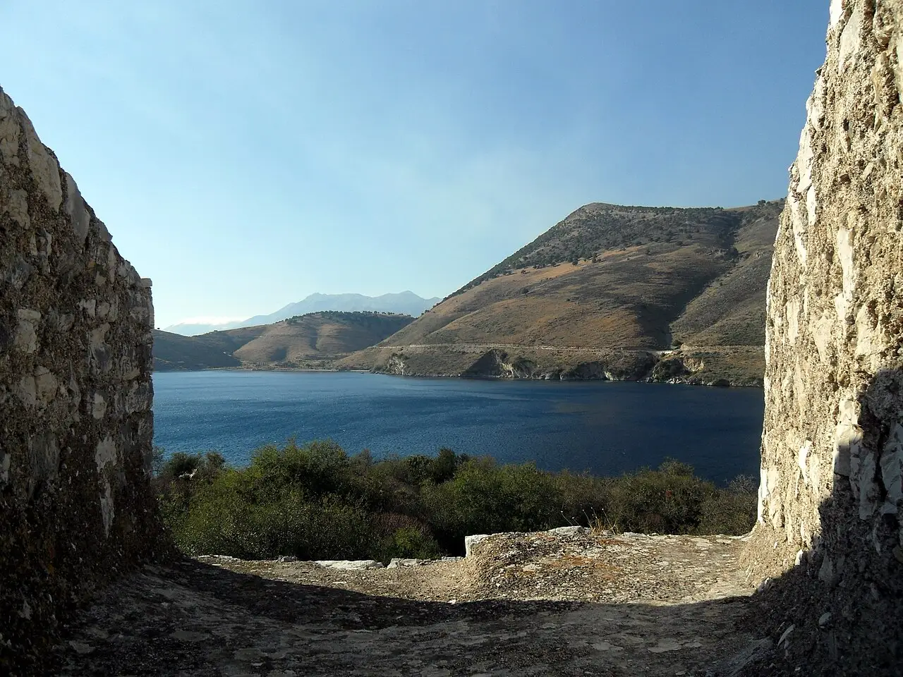 The blue water of Porto Palermo bay and the bare Ceraunian hills framed by two stretches of fortress wall