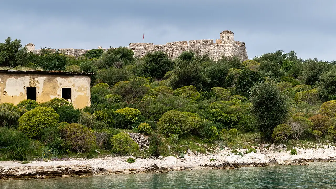 Porto Palermo Castle on its wooded headland seen from the water, with a derelict yellow building on the shore below