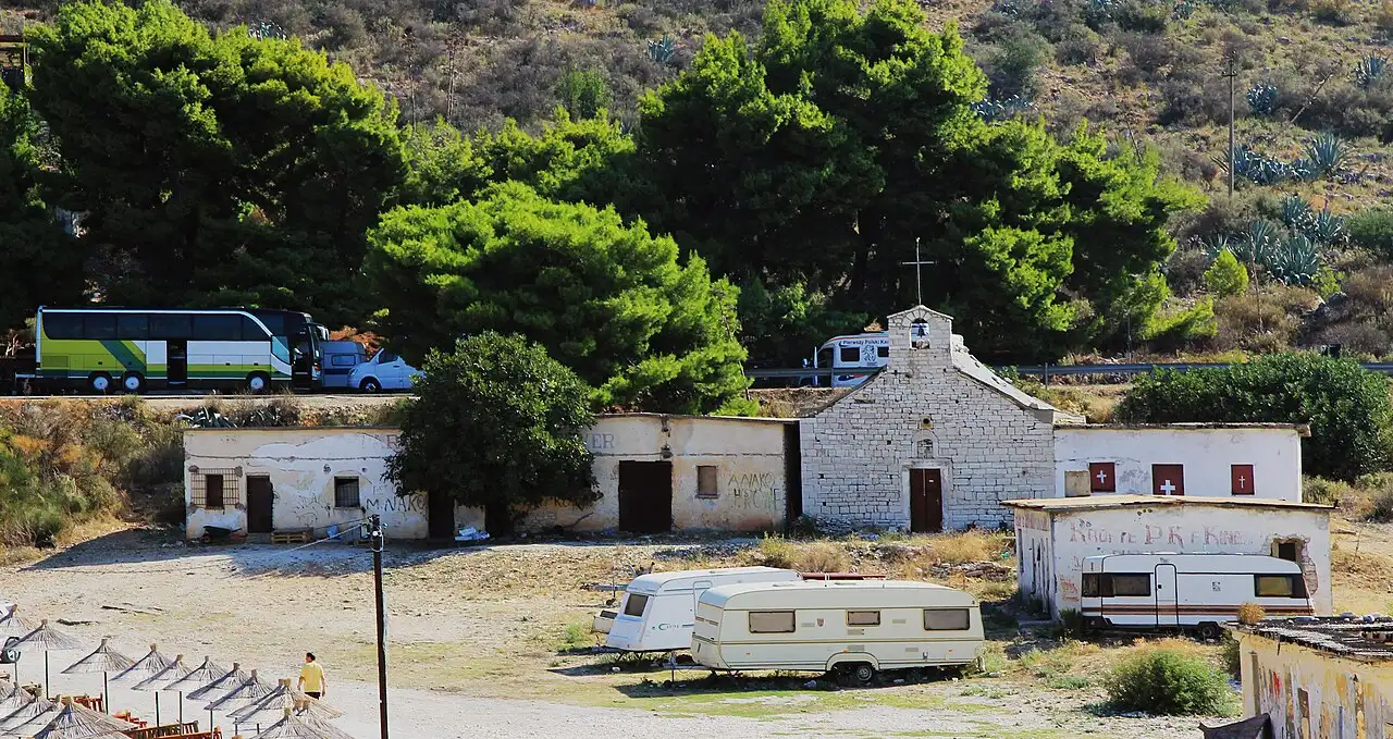 A small white chapel with a bell gable standing between abandoned barracks and parked caravans at Porto Palermo