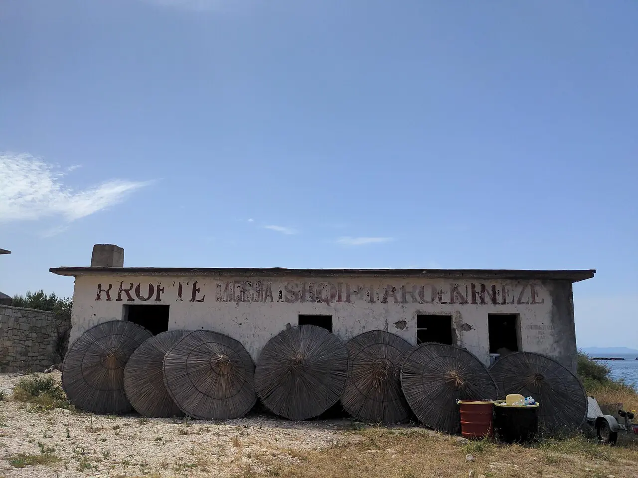 A derelict single-storey concrete building at Porto Palermo painted with a faded red slogan, straw beach parasols stacked against its wall
