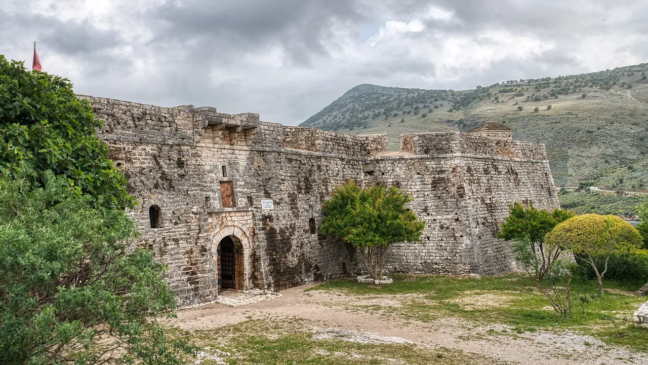 The outer wall and arched entrance of Porto Palermo Castle with the Albanian flag flying above the rampart