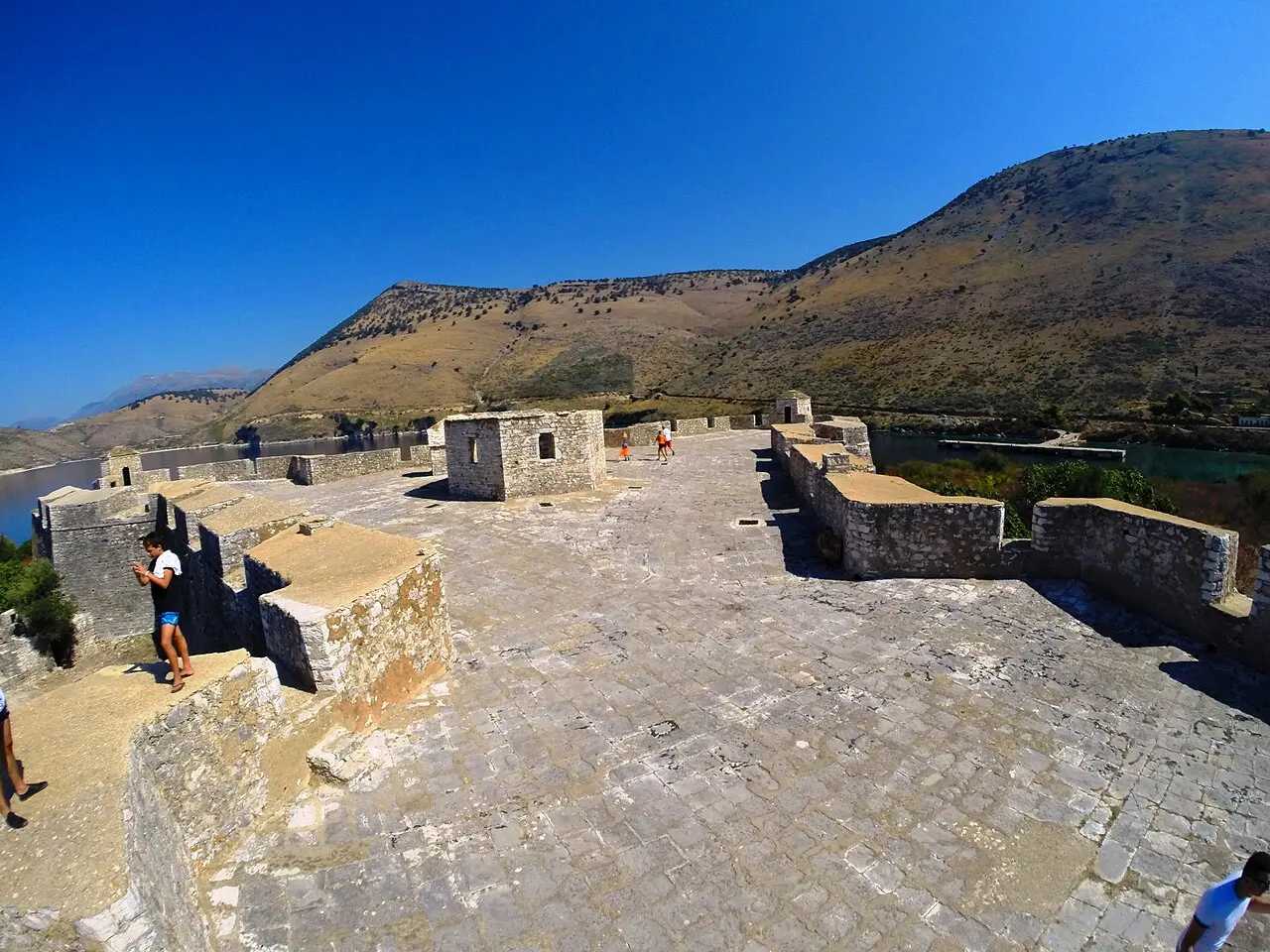 The stone roof terrace of Porto Palermo Castle with low walls, guard houses and a few visitors, the bay and dry hills behind