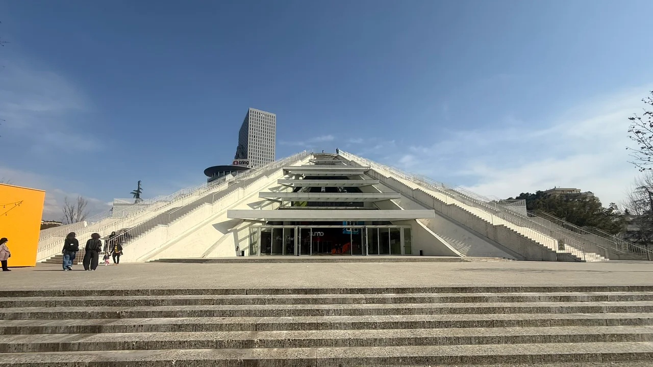 The white renovated Pyramid of Tirana seen head-on from the plaza, with staircases running up its sloping beams, a glass entrance at the base and a yellow pavilion on the left