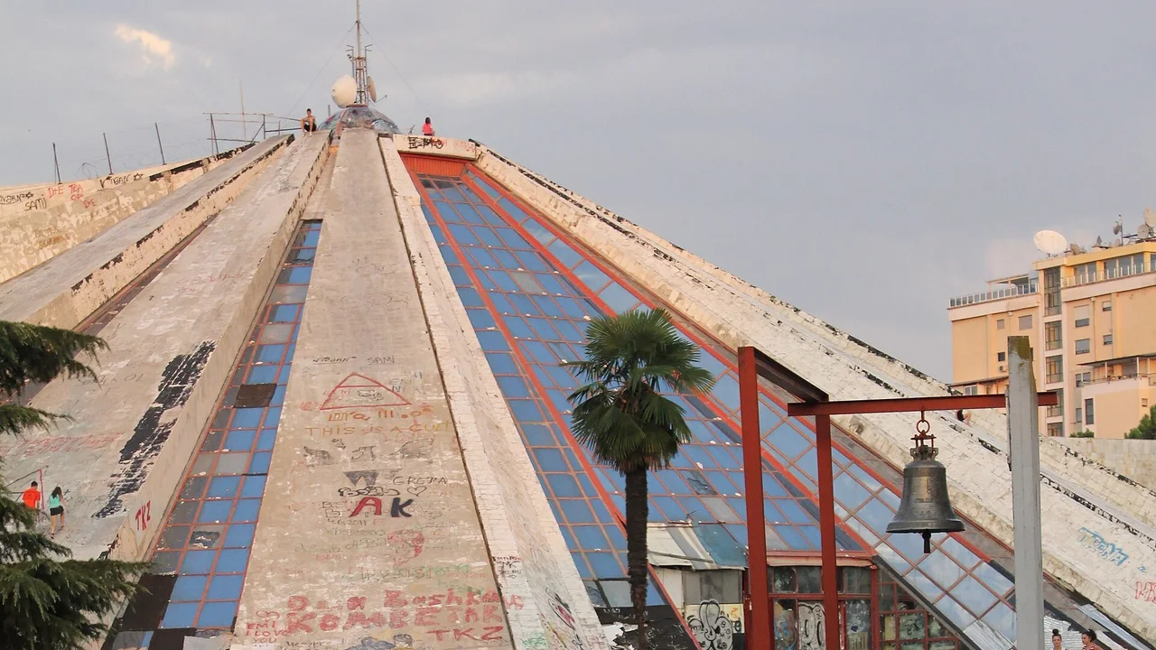 The Pyramid of Tirana in July 2012, its bare concrete beams covered in graffiti between strips of blue glass, two people climbing a beam on the left and two sitting at the top, a palm tree and a bronze bell on a red frame in front
