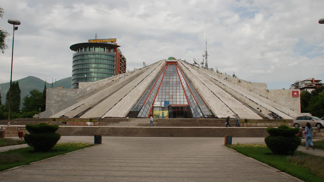 The Pyramid of Tirana before renovation, with weathered grey concrete beams, a red-framed glass atrium, satellite dishes on top and a round office tower behind