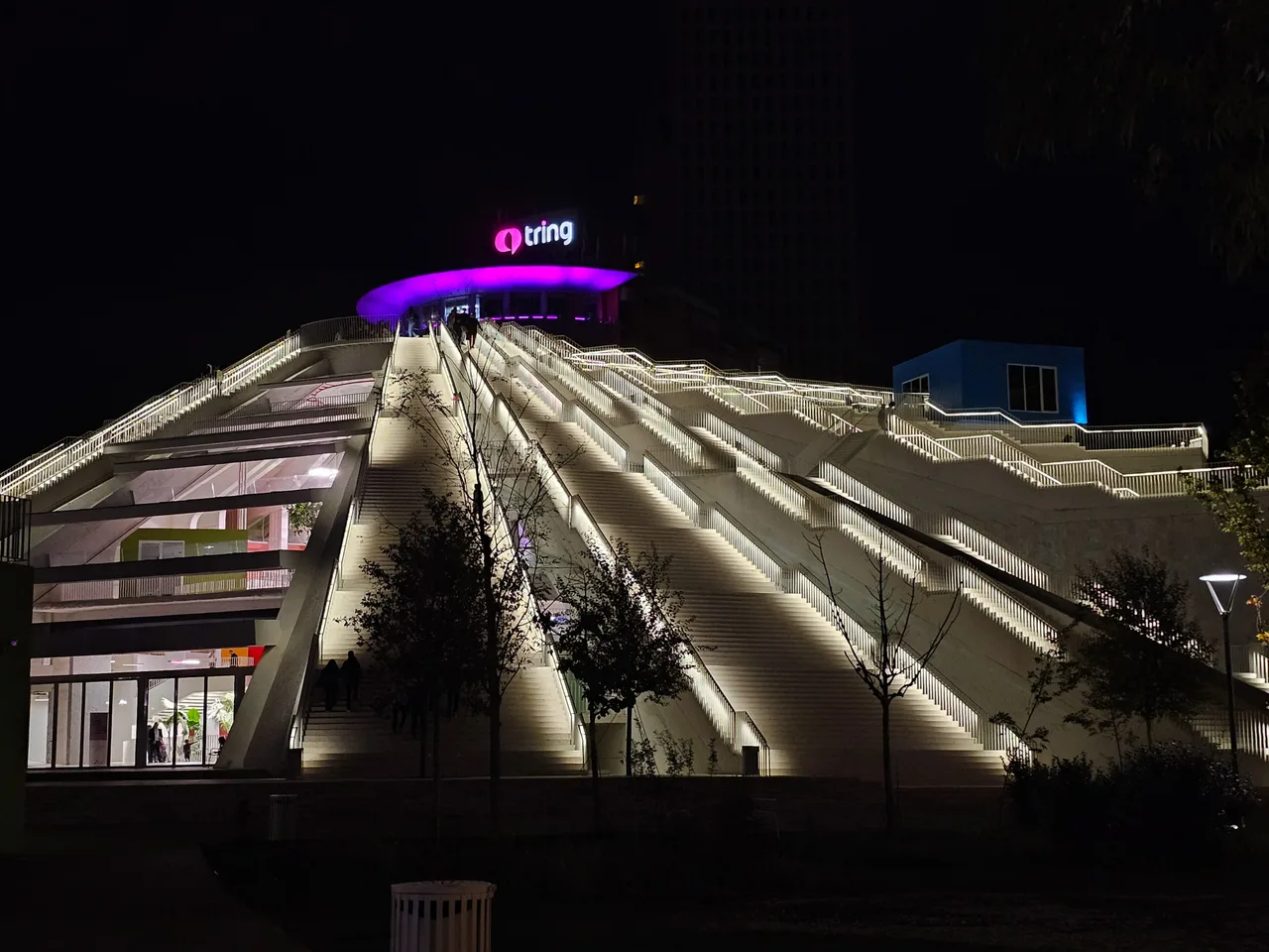 The Pyramid of Tirana at night with white light strips along its staircases, people climbing, and a glowing purple Tring sign at the top