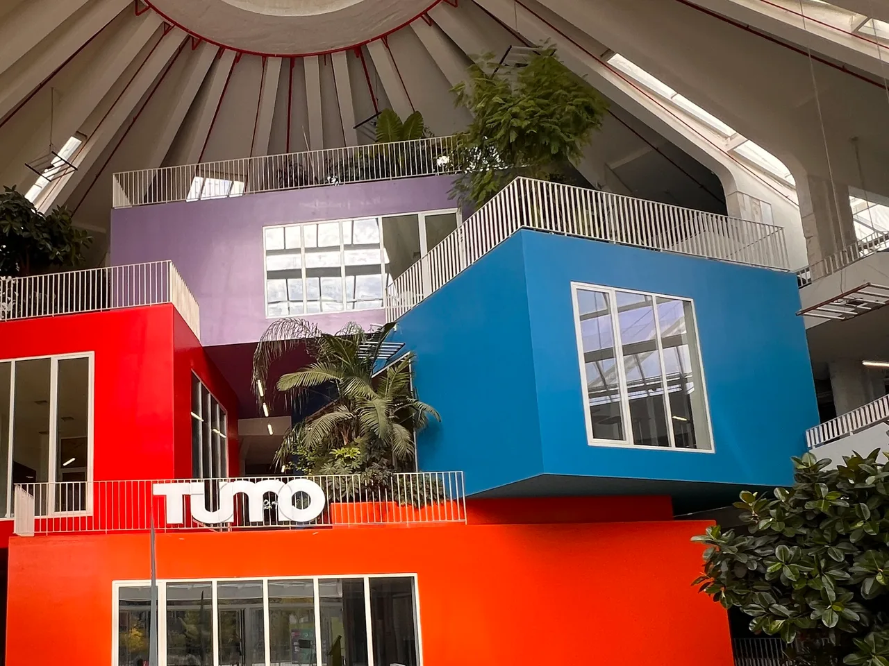Stacked red, blue and purple boxes with white railings and plants under the ribbed concrete roof inside the Pyramid, with a TUMO sign on the front