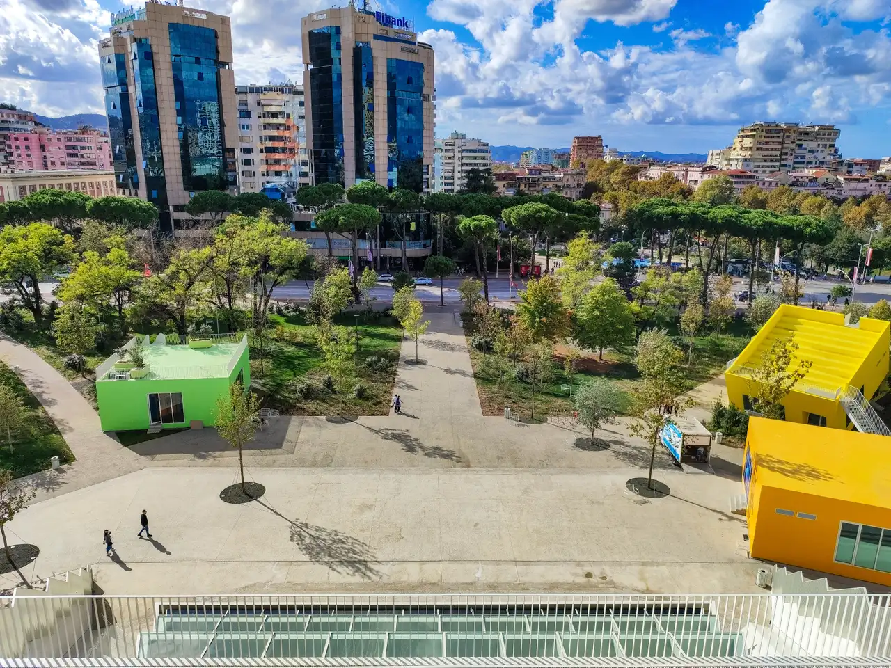 View from high on the Pyramid over a paved plaza with green and yellow boxes, a park of young trees, the boulevard and two glass office towers under a cloudy blue sky