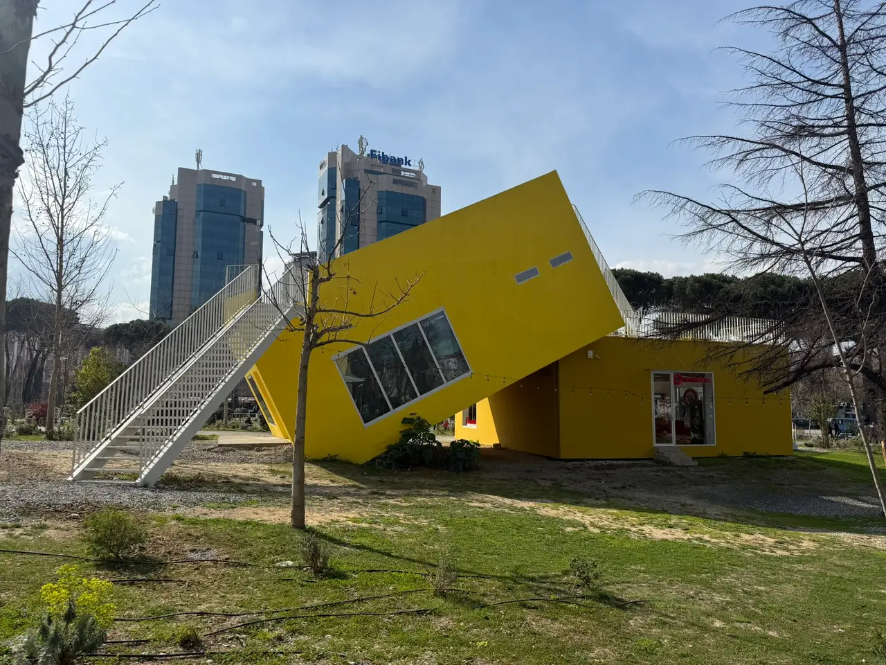 A bright yellow box pavilion tilted at an angle on a lawn next to the Pyramid, with a white external staircase and two glass office towers behind