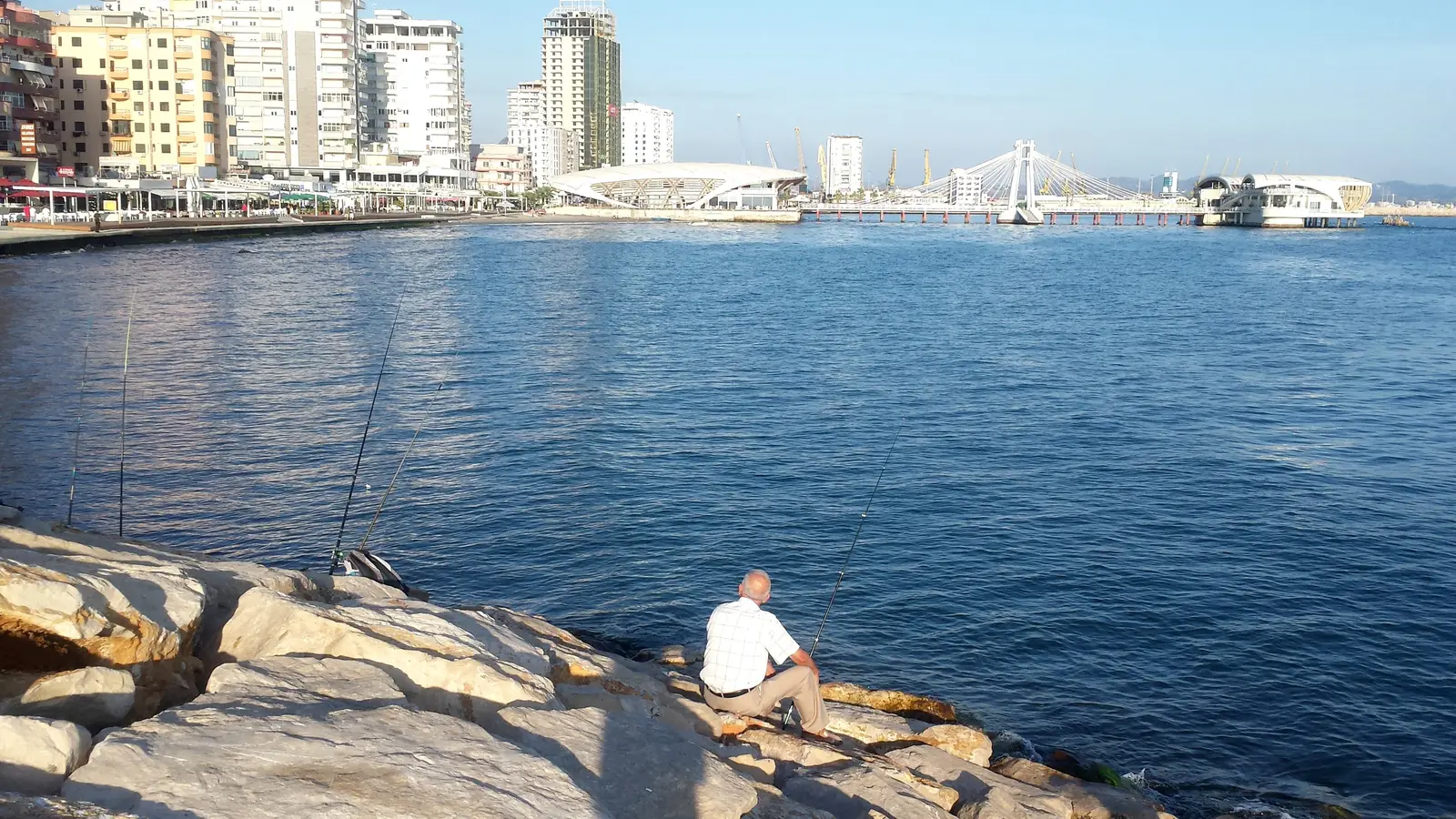 An older man in a white checked shirt sits fishing on the rocks of the Durrës seafront, with calm blue water, apartment towers and the white pier pavilions behind
