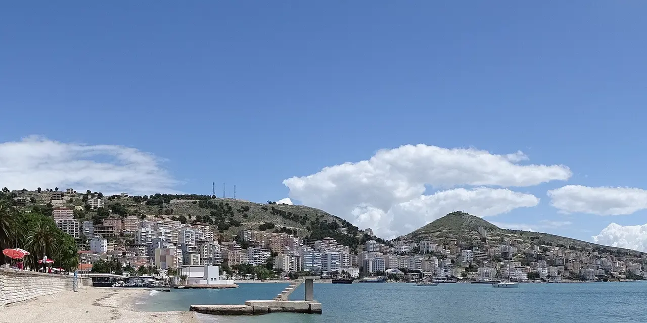 Sarandë's seafront with a low stone wall, palm trees and a promenade curving round the bay to apartment blocks climbing a hillside
