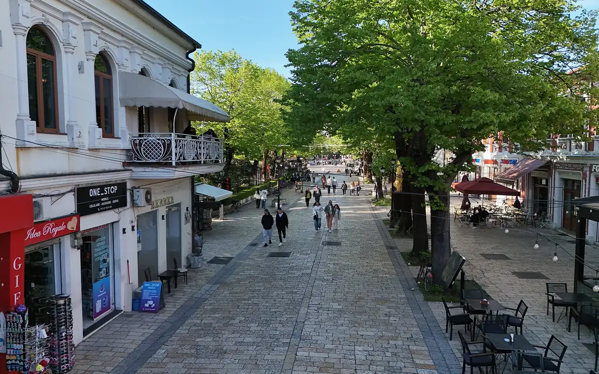 A pedestrian street in Shkodër lined with two-storey houses, café tables and tall leafy trees, with a few people walking on the paving