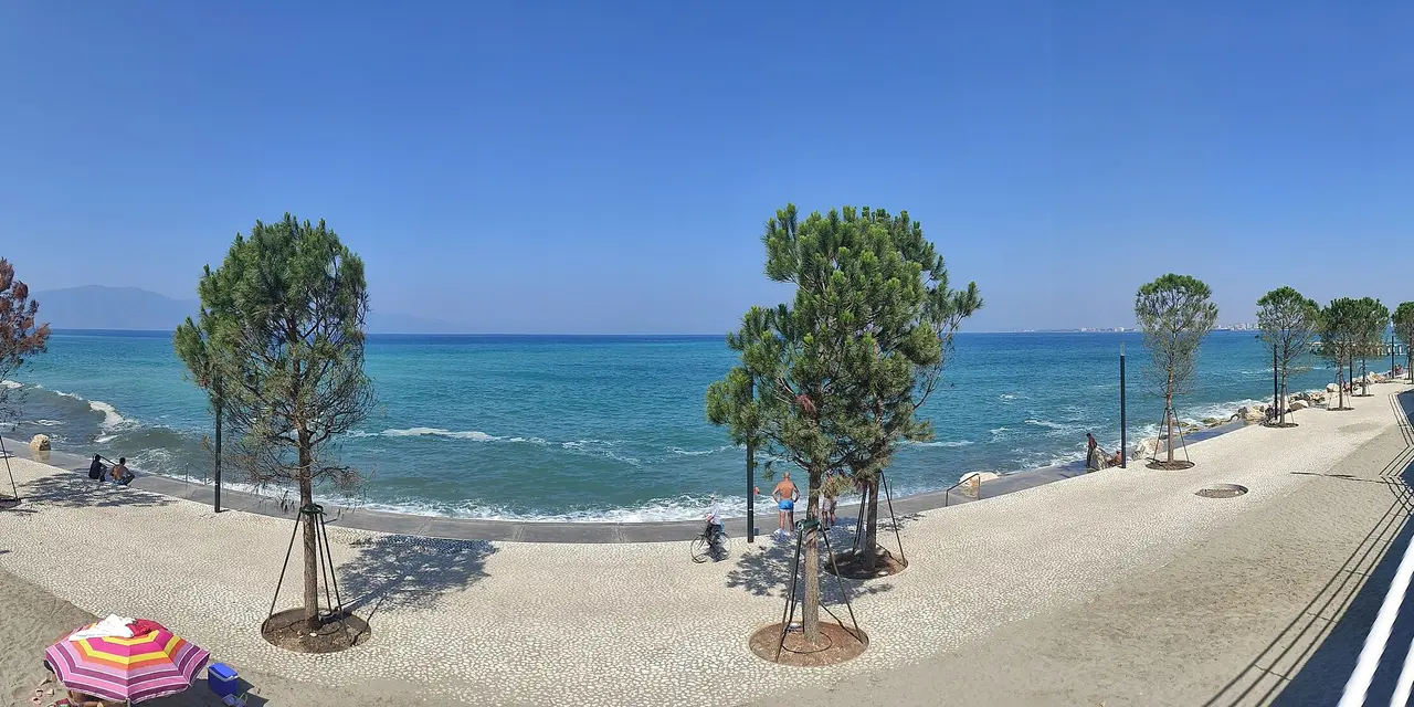 The Lungomare in Vlorë: a pale paved promenade with young pines curving along a turquoise sea, swimmers on the shore and mountains across the bay