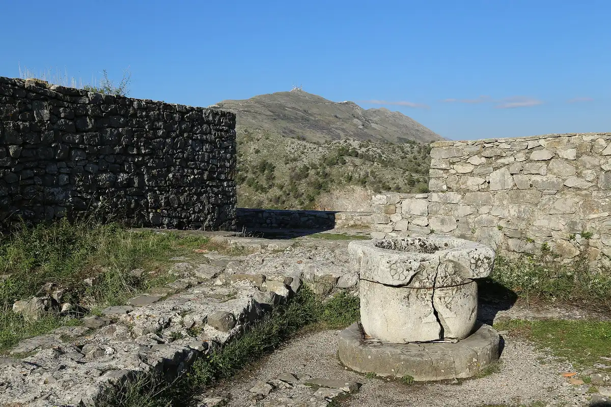 A carved stone cistern head in a courtyard of Rozafa Castle, with walls and a mountain behind