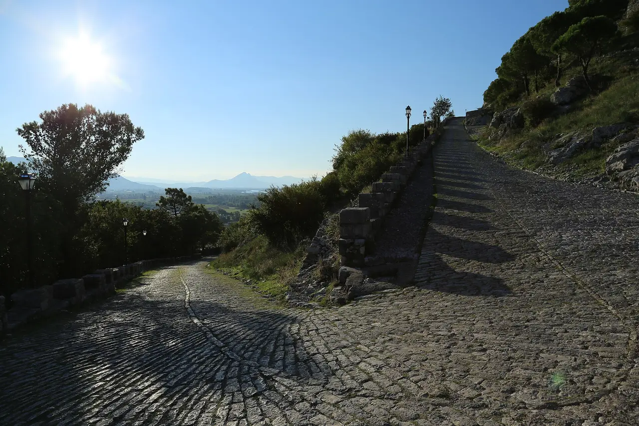 The steep cobbled road climbing toward Rozafa Castle, with lamps along the verge and mountains across the valley