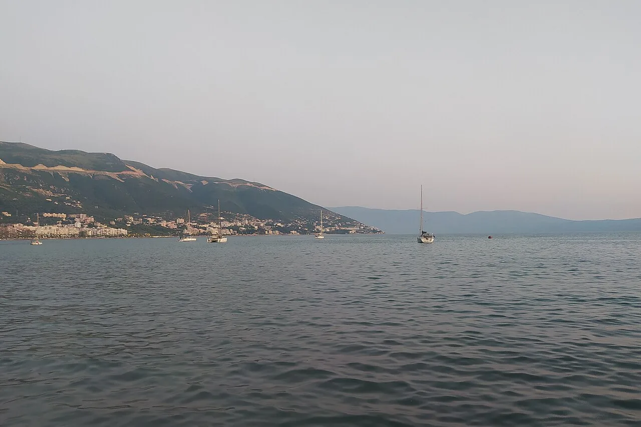 The city of Vlora curving around its bay under the mountains, with sailboats moored offshore in the evening light