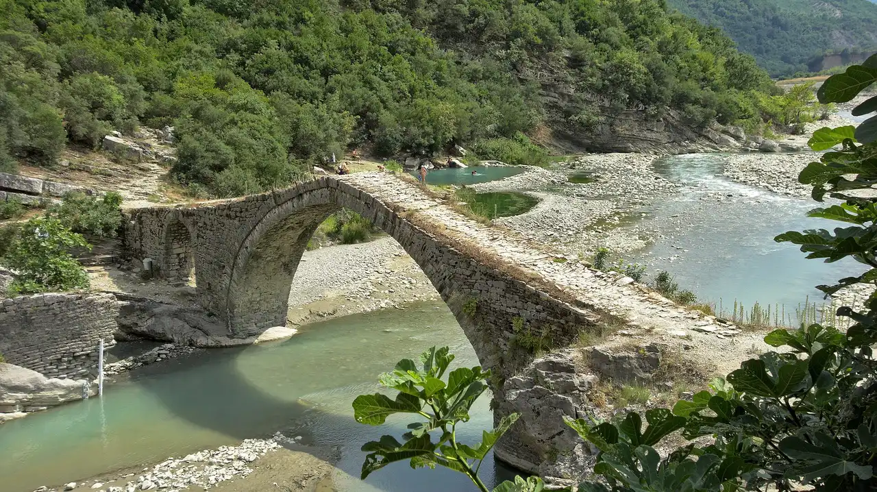 The arched Ottoman Kadiu Bridge over the turquoise Lengarica river at the Benje thermal baths near Permet, green canyon walls behind