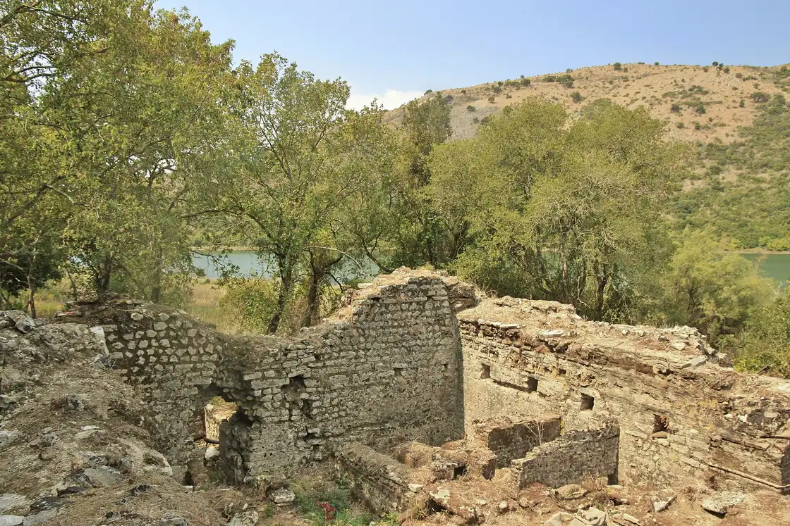 Stone walls and ruined buildings of ancient Butrint among trees, with the lagoon and wooded hills behind