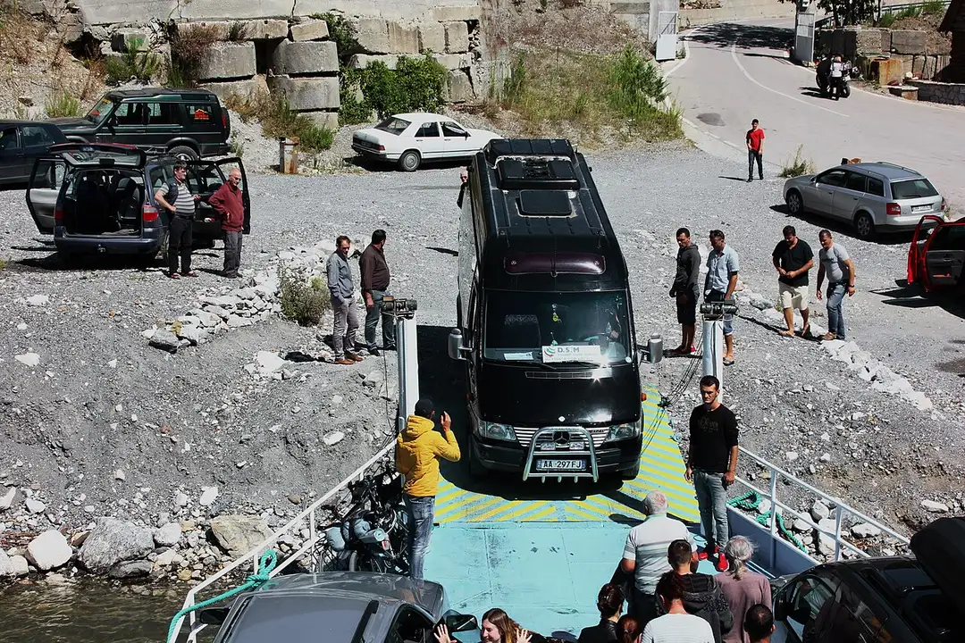 Cars and a minibus queued to drive off a vehicle ferry onto a gravel landing beside Lake Koman