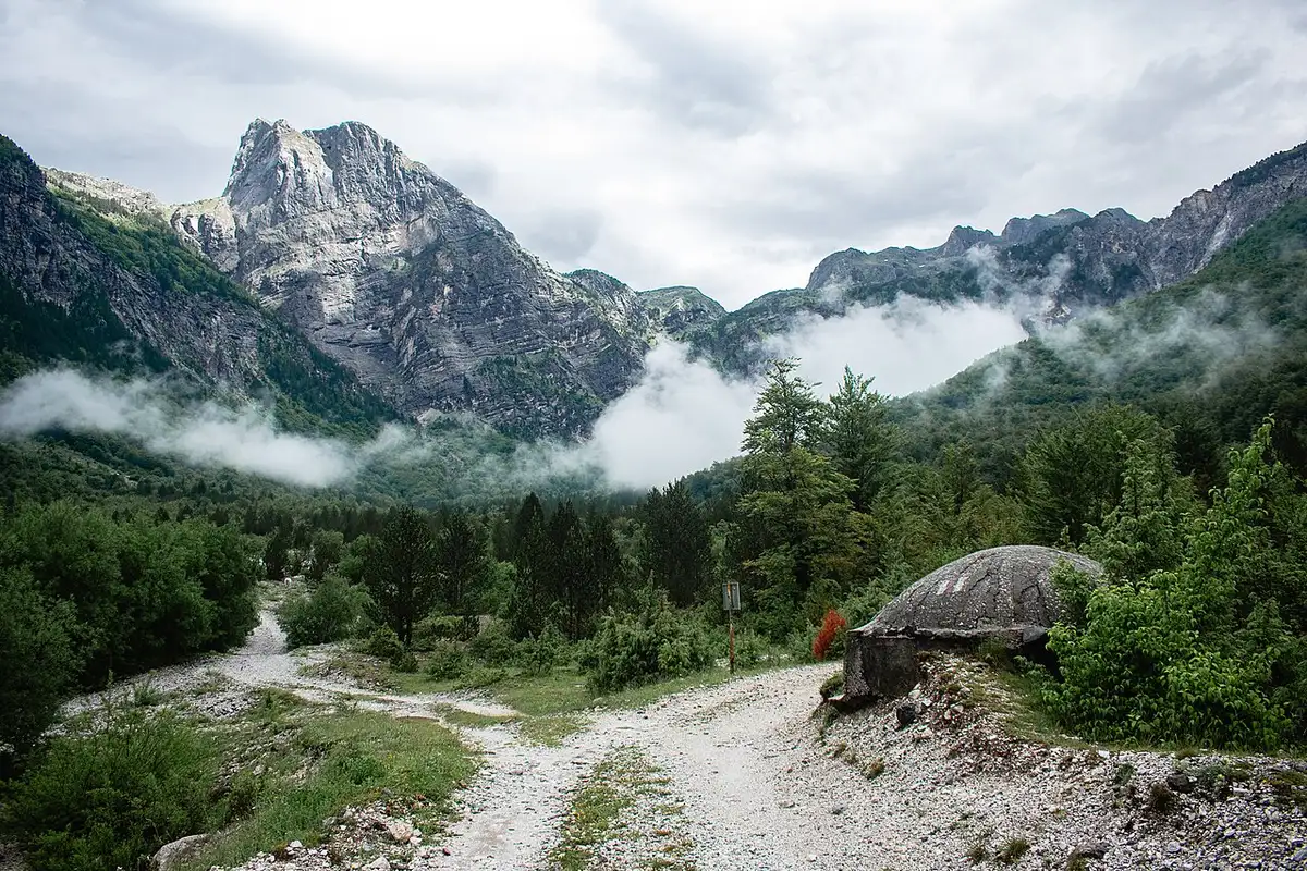 A concrete Hoxha-era bunker beside a gravel trail in Theth National Park, with clouds drifting around the peaks behind