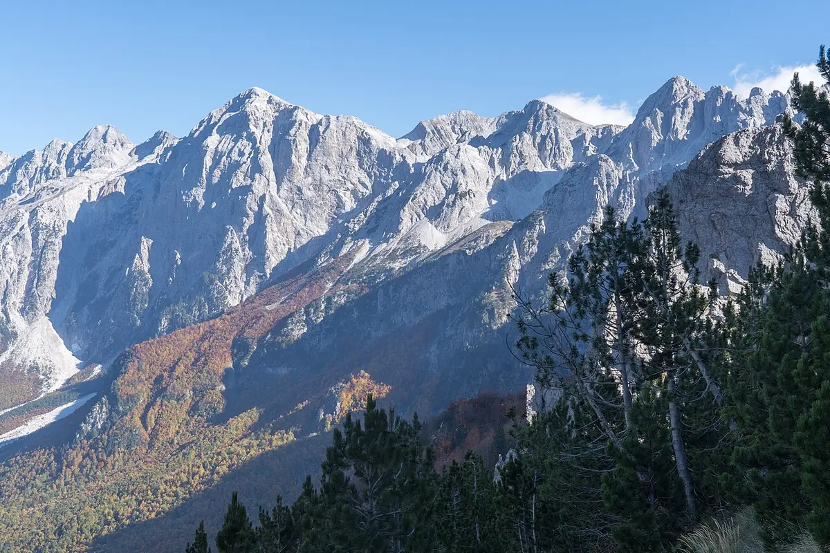 Bare limestone peaks and a forested slope above the open floor of Valbona Valley in autumn
