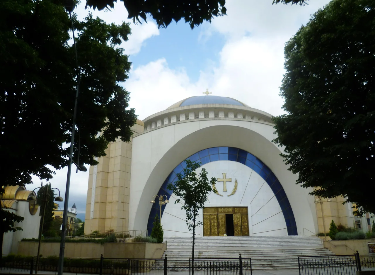 The white and blue arched facade of the Resurrection of Christ Orthodox Cathedral in Tirana, framed by trees