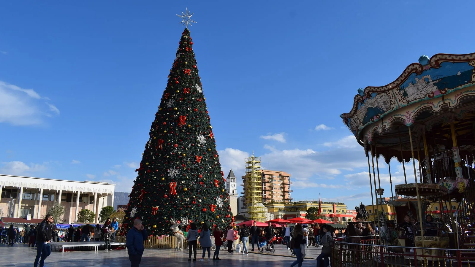 The giant decorated Christmas tree on Skanderbeg Square in Tirana beside a vintage carousel, with the clock tower behind