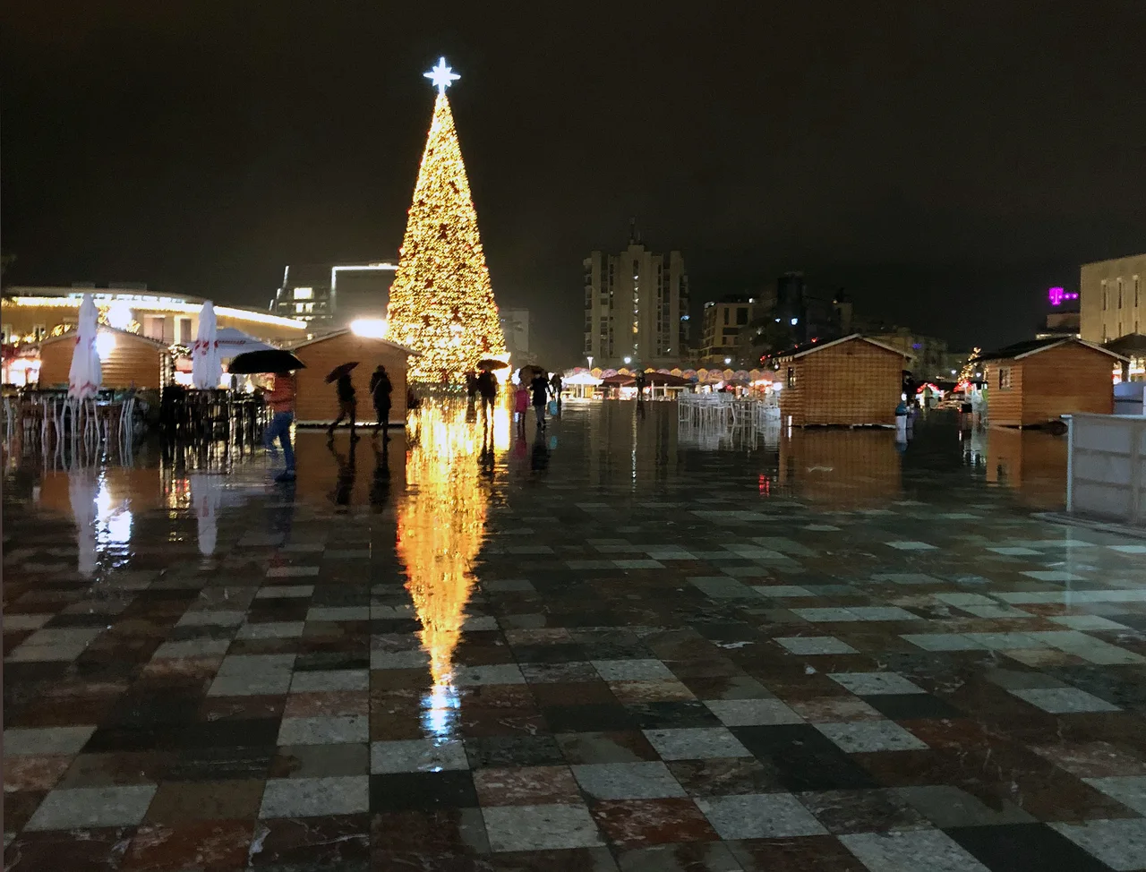 The lit Christmas tree reflected in the wet paving of Skanderbeg Square at night, with wooden market huts and people under umbrellas