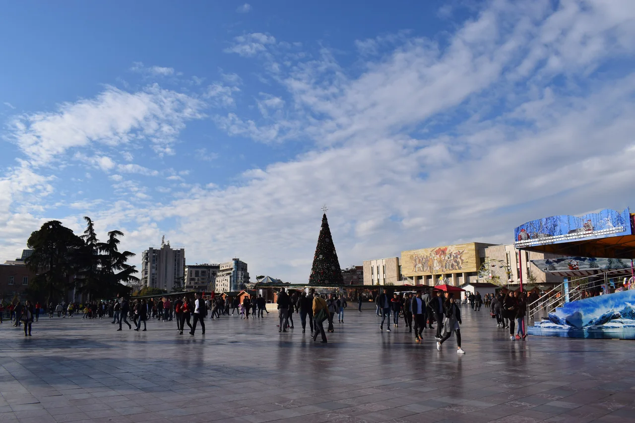 Crowds crossing Skanderbeg Square on a sunny afternoon in November 2018, with the Christmas tree, market huts and the National History Museum mosaic in the distance