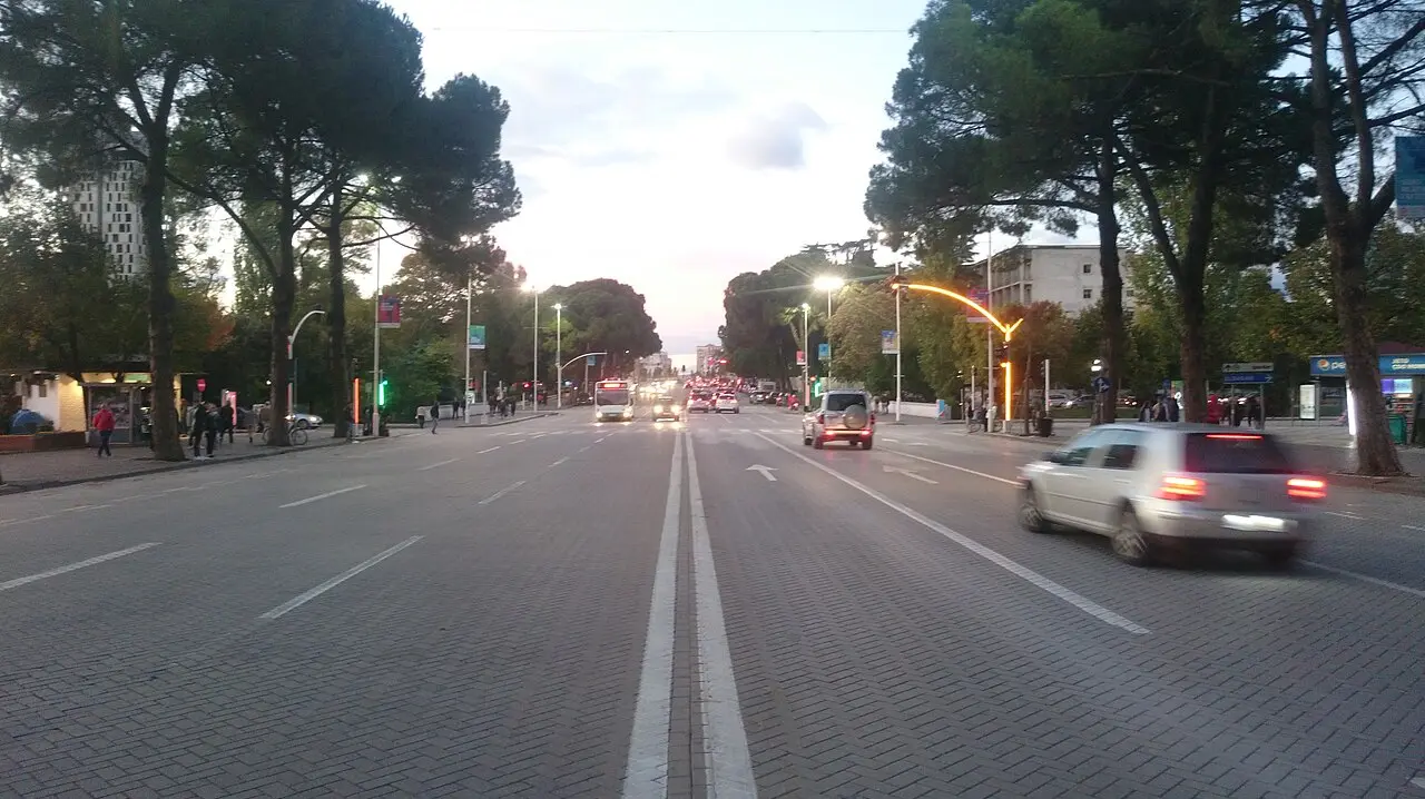 The main Deshmoret e Kombit boulevard in central Tirana at dusk, tree-lined with traffic and modern buildings