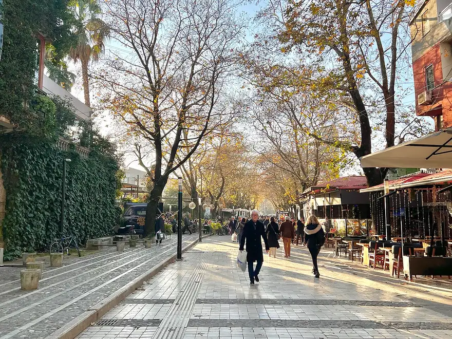 A leafy pedestrian street in central Tirana with people walking and cafe terraces with fairy lights